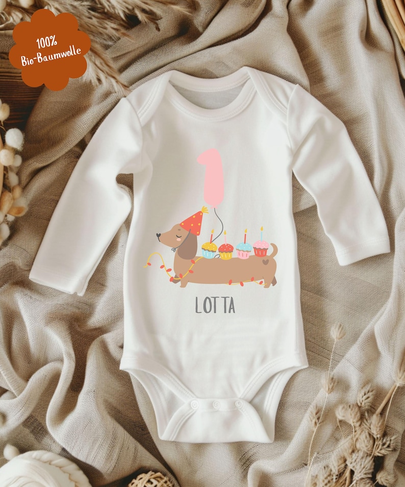 Op de afbeelding: Witte babybody met lange mouwen met een illustratie van een teckel met een feesthoed, cupcakes en een roze ballon. De tekst "LOTTA" staat eronder.