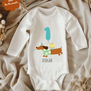 Op de afbeelding: Witte babybody met lange mouwen, met een cartoon teckel met een feesthoed en een groene trui. De teckel houdt een toeter vast en staat naast een verjaardagstaart en een blauwe ballon met het nummer 1. De naam "OSKAR" staat eronder gedrukt.