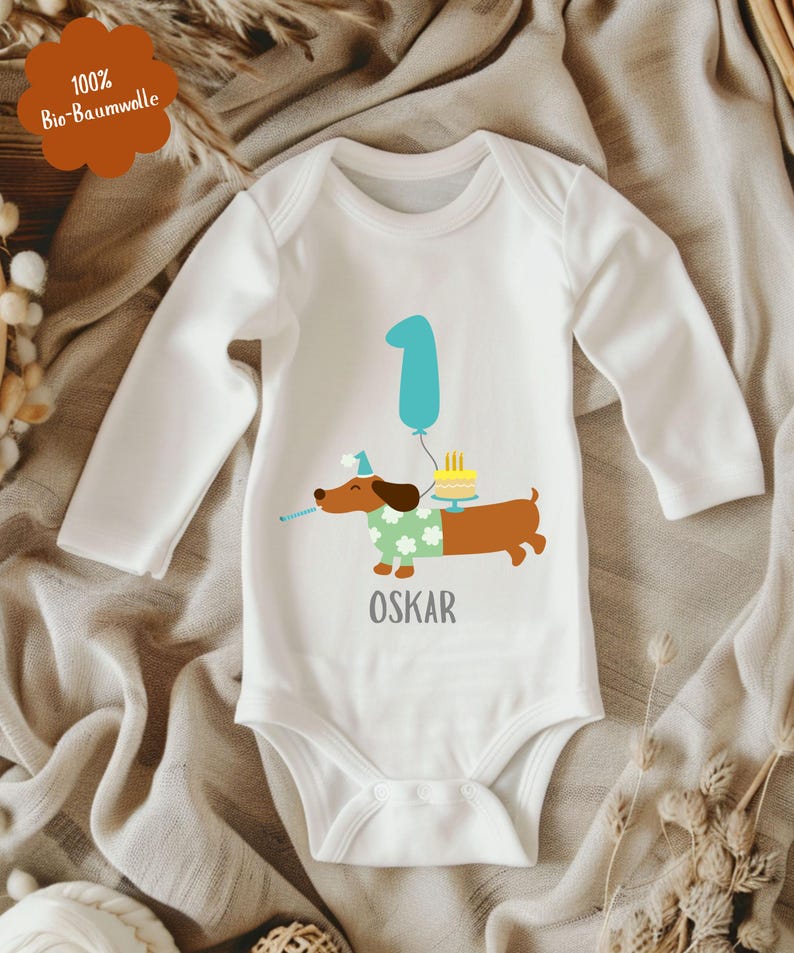 Op de afbeelding: Witte babybody met lange mouwen, met een cartoon teckel met een feesthoed en een groene trui. De teckel houdt een toeter vast en staat naast een verjaardagstaart en een blauwe ballon met het nummer 1. De naam "OSKAR" staat eronder gedrukt.