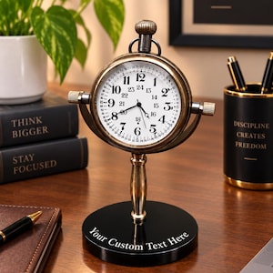 May include: A brass-toned desk clock with a white face featuring both 12-hour and 24-hour markings, mounted on a vertical stand with a circular black base. The base displays the white text "Your Custom Text Here" in a serif font.