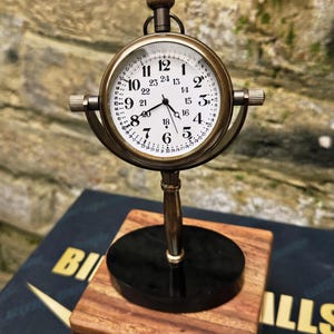 Op de afbeelding: Een zakhorloge in vintage-stijl op een standaard. De witte wijzerplaat heeft zwarte cijfers en wijzers, met een messingkleurig frame. Het staat op een zwarte basis en een houten blok. De klok is ongeveer 7.6 cm hoog.