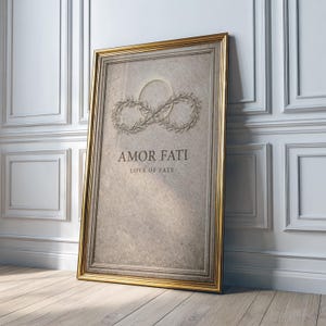 May include: A framed artwork with a gold-coloured frame, leaning against a white wall. The artwork features the words "AMOR FATI LOVE OF FATE" and an infinity symbol design with a circular element and leafy branches.