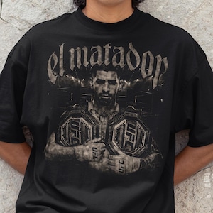 May include: Black t-shirt featuring a sepia-toned image of a UFC fighter holding championship belts. The text "el matador" is arched above the fighter's image. The person in the image is wearing a black baseball cap.