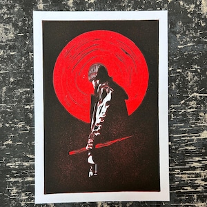 May include: A black and red linocut print featuring a silhouetted figure in a beanie and coat, holding a long object, set against a large red circle. The print is on white paper with a black background.