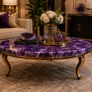 May include: A round coffee table featuring a polished purple amethyst gemstone tabletop surface and curved gold metal legs. The table is decorated with a black Chanel book, a purple amethyst cluster, and a glass vase filled with white flowers.
