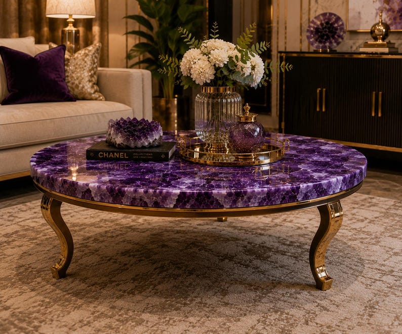 May include: A round coffee table featuring a polished purple amethyst gemstone tabletop surface and curved gold metal legs. The table is decorated with a black Chanel book, a purple amethyst cluster, and a glass vase filled with white flowers.