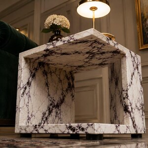 May include: A square marble side table with a white and dark gray veined pattern. The table has a hollow center and sits on four black feet. A lamp and a vase of flowers are on top of the table.