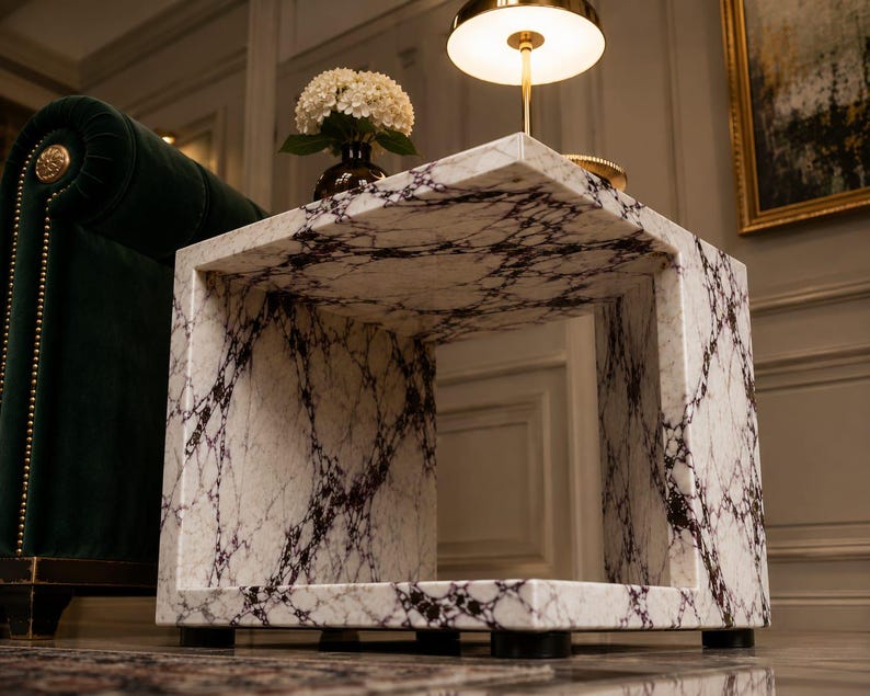 May include: A square marble side table with a white and dark gray veined pattern. The table has a hollow center and sits on four black feet. A lamp and a vase of flowers are on top of the table.