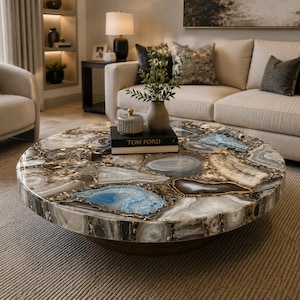 May include: A round coffee table featuring a tabletop composed of numerous polished agate slices in shades of blue, gray, white, and brown. The surface has a glossy finish, and a black book titled Tom Ford sits in the center with a small vase.