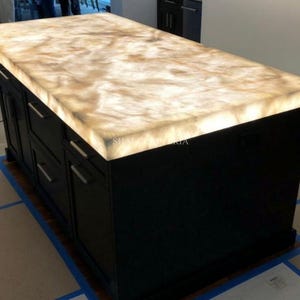 May include: A large, black kitchen island with a glowing, light-colored countertop. The countertop has a marbled appearance with shades of beige and brown. The island features multiple drawers and cabinet doors with silver handles. The text "SHAI CRAFTERIA" is visible.