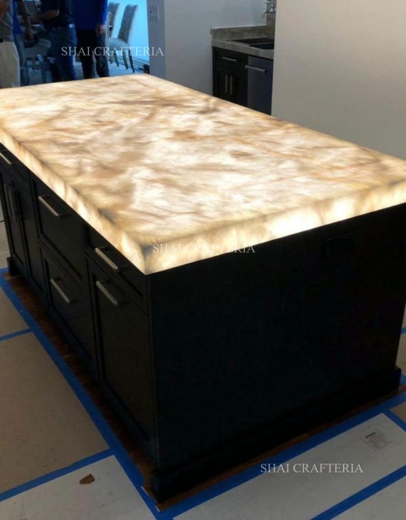 May include: A large, black kitchen island with a glowing, light-colored countertop. The countertop has a marbled appearance with shades of beige and brown. The island features multiple drawers and cabinet doors with silver handles. The text "SHAI CRAFTERIA" is visible.
