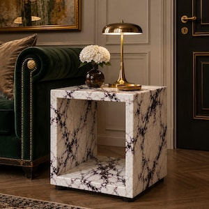 May include: A white marble side table with dark purple veining. A gold-toned lamp with a domed shade sits on top, next to a small vase of white flowers. The table is square with an open center.