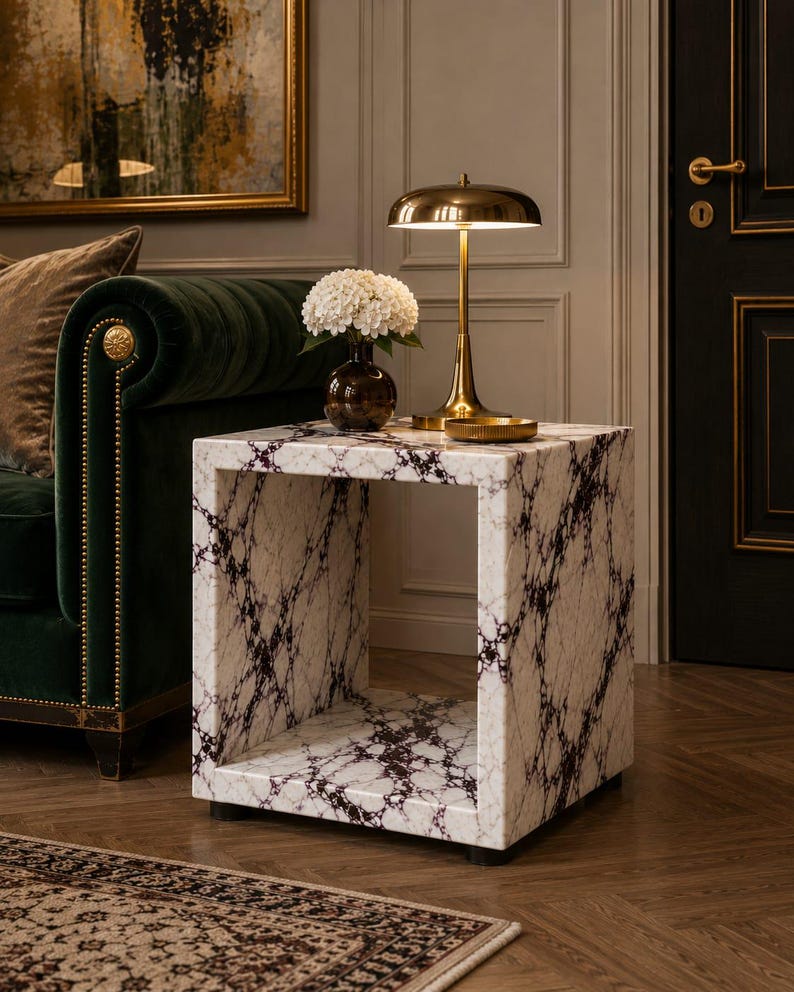 May include: A white marble side table with dark purple veining. A gold-toned lamp with a domed shade sits on top, next to a small vase of white flowers. The table is square with an open center.