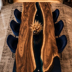 May include: A rectangular dining table featuring a live-edge wood slab with a central dark blue resin river inlay, surrounded by six navy blue upholstered chairs. A vase containing dried pampas grass sits in the centre of the table surface.