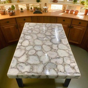 May include: A rectangular table with a white and gray stone top. The stone has a pattern of irregular, rounded shapes. The table has black legs and is set in a kitchen with wooden cabinets and a window.