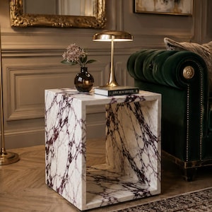 May include: A white marble side table with deep purple veining. A gold-toned lamp, a small vase with flowers, and a book are on the table. The table is in a room with a green velvet sofa and a patterned rug.