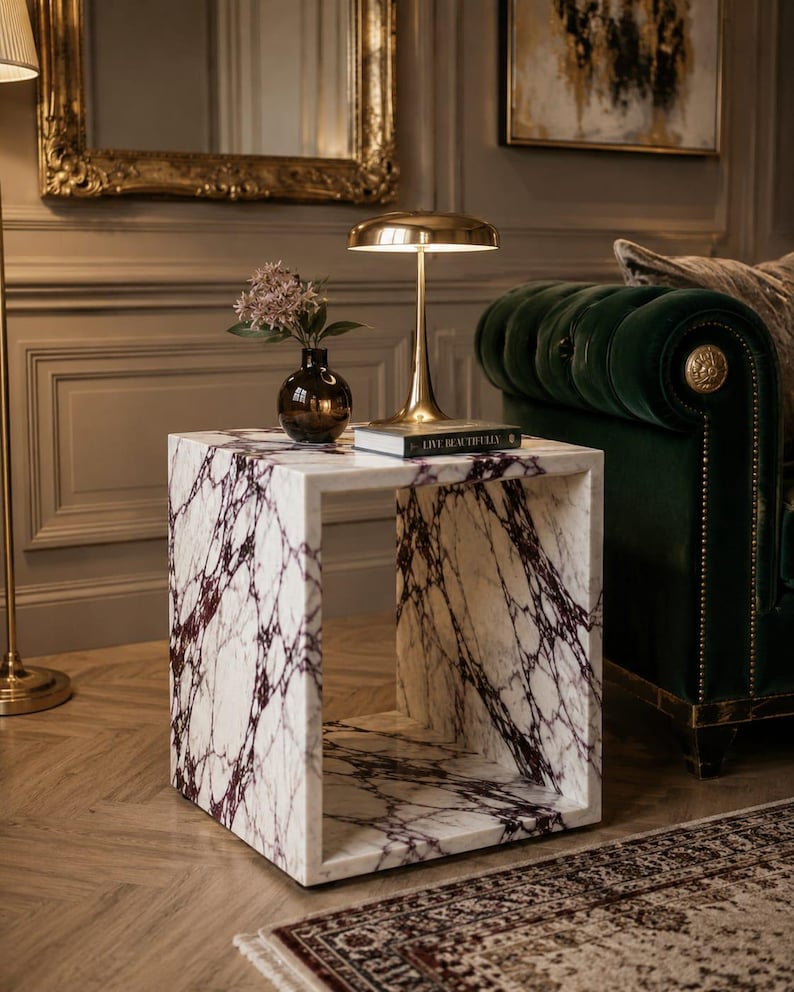 May include: A white marble side table with deep purple veining. A gold-toned lamp, a small vase with flowers, and a book are on the table. The table is in a room with a green velvet sofa and a patterned rug.