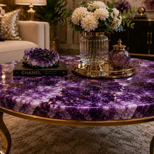 May include: A round coffee table featuring a polished purple amethyst gemstone surface and curved gold metal legs. On the table sits a black Chanel book, a large amethyst geode, a gold tray, a glass vase with white flowers, and a purple jar.