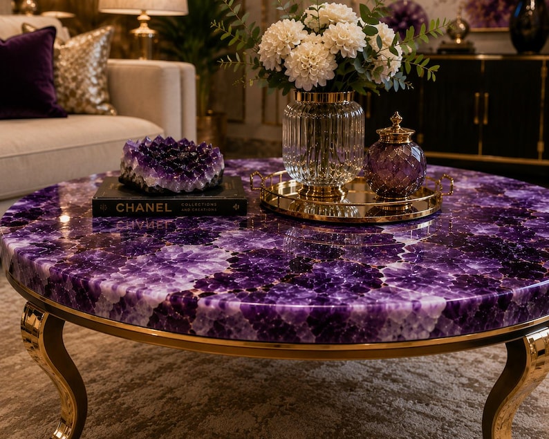 May include: A round coffee table featuring a polished purple amethyst gemstone surface and curved gold metal legs. On the table sits a black Chanel book, a large amethyst geode, a gold tray, a glass vase with white flowers, and a purple jar.