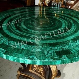 Puede incluir: Mesa redonda de malaquita con un llamativo patrón verde y negro. La tapa de la mesa presenta círculos concéntricos de malaquita, y la base tiene un diseño dorado y negro. El texto "SHAI CRAFTERIA" está impreso en la mesa.