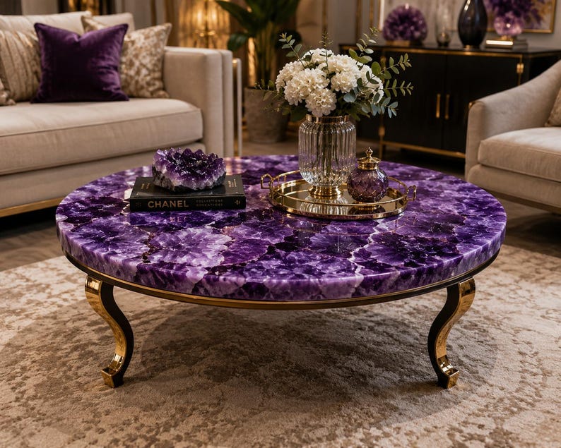 May include: A round coffee table featuring a deep purple amethyst gemstone surface with a polished finish. The table is supported by four curved gold-toned metal legs. A black book titled CHANEL, a purple crystal cluster, and a gold tray are on top.
