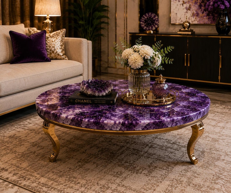 May include: A round coffee table featuring a polished purple amethyst gemstone surface and curved gold metal legs. The table is topped with a black book labeled CHANEL, a large amethyst geode cluster, a glass vase with white flowers, and a gold tray.