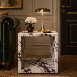 May include: A marble cube side table with a white and burgundy veined pattern. A brass desk lamp and a vase of white flowers sit on top. The lamp has a rounded shade and a slender stem. The room has a dark green sofa and a patterned rug.