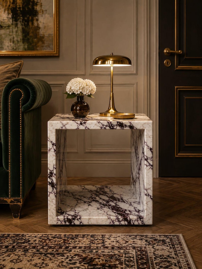 May include: A marble cube side table with a white and burgundy veined pattern. A brass desk lamp and a vase of white flowers sit on top. The lamp has a rounded shade and a slender stem. The room has a dark green sofa and a patterned rug.