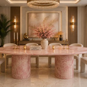 May include: A luxurious dining room featuring a pink oval table with fluted columns, surrounded by cream-colored chairs. A large floral arrangement sits atop the table. The room is illuminated by a chandelier and wall sconces.