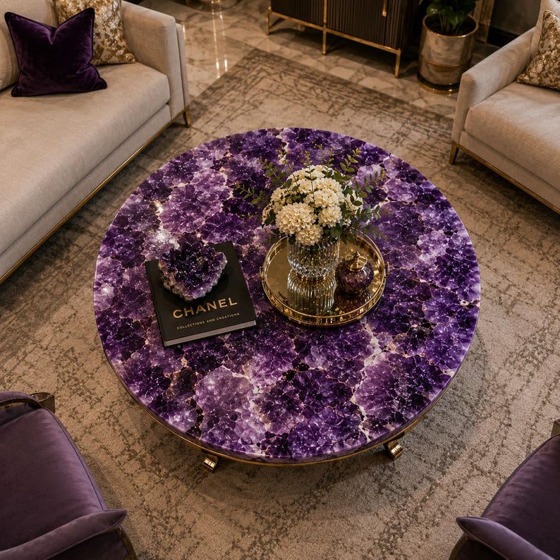 May include: A round coffee table featuring a surface made of deep purple amethyst crystal clusters. On the table sits a black book titled CHANEL COLLECTIONS AND CREATIONS, a small amethyst cluster, and a gold tray holding a glass vase with white flowers.