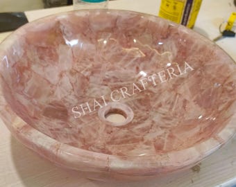 May include: A round, pink stone sink with a polished finish. The sink has a central drain hole and the text "SHAI CRAFTERIA" is visible on the surface. The sink is made of a mosaic of pink and white stone.