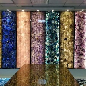 May include: A series of vertical rectangular panels featuring backlit semi-precious stone slabs in various colors including gold, blue, peach, purple, and teal, arranged side-by-side, with a polished brown stone surface in the foreground.