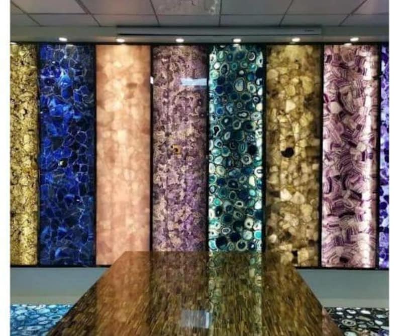 May include: A series of vertical rectangular panels featuring backlit semi-precious stone slabs in various colors including gold, blue, peach, purple, and teal, arranged side-by-side, with a polished brown stone surface in the foreground.
