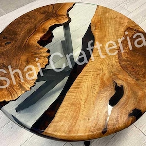 May include: A round coffee table featuring two natural-edged wood slabs separated by a clear resin river inlay. The wood displays a rich brown grain with dark knots. The text Shai Crafteria is overlaid on the surface. It has a black metal base.