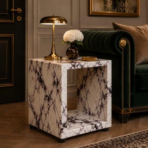 May include: A marble-patterned side table with a square design. The table features a white base with black veining. A gold-toned lamp and a vase with white flowers sit on top. The table is in a room with a green sofa and a patterned rug.