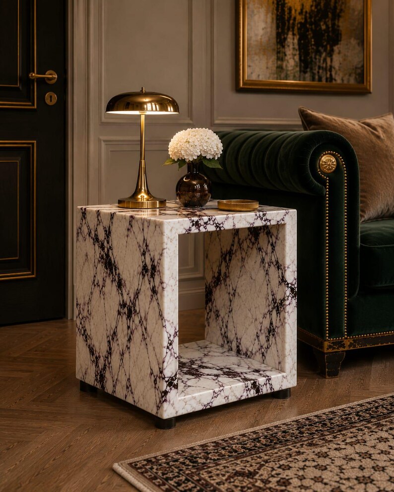May include: A marble-patterned side table with a square design. The table features a white base with black veining. A gold-toned lamp and a vase with white flowers sit on top. The table is in a room with a green sofa and a patterned rug.
