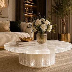 Pode incluir: Uma mesa de centro redonda e branca com base e tampo texturizados. A mesa está numa sala de estar, com um sofá, detalhes dourados e um vaso de flores brancas. A mesa tem cerca de 100 cm de diâmetro.