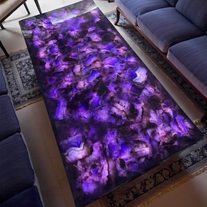 Può includere: Un tavolino rettangolare con superficie in ametista lucida. La superficie del tavolo è un mosaico di tonalità viola intenso, lavanda e bianche, creando un effetto visivo sorprendente. Il tavolo è circondato da divani blu e un tappeto fantasia.