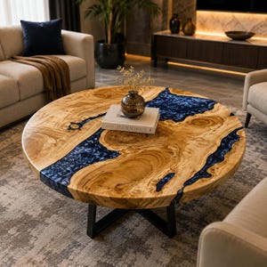 May include: A round coffee table featuring a natural light wood grain surface with a prominent, flowing inlay of deep blue resin. The table is supported by a black metal cross-base and holds a small glass vase with dried flowers and a book.