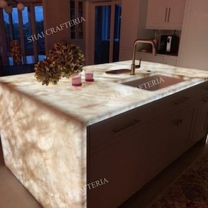 May include: A backlit kitchen island with a marble countertop and a built-in sink. The island has a light brown cabinet base with a gold faucet. The countertop has a natural stone pattern. The text "SHAI CRAFTERIA" is visible.