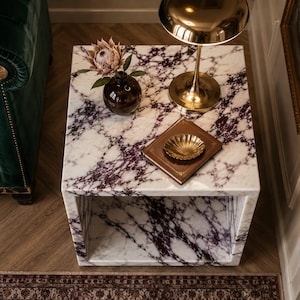 Könnte beinhalten: Ein quadratischer Beistelltisch aus Marmor mit weißer Basis und lila Maserung. Eine kleine braune Vase mit einer Blume, eine goldene Lampe und eine dekorative goldene Schale auf einem braunen Buch befinden sich auf dem Tisch. Der Tisch steht in einem Raum mit einem grünen Sofa.