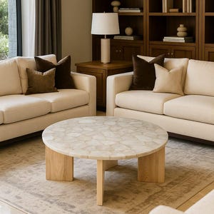 May include: A round coffee table featuring a mosaic-style top composed of irregular, light-colored stone pieces. The table is supported by three thick, light-toned wooden legs arranged in a tripod configuration, providing a stable base.