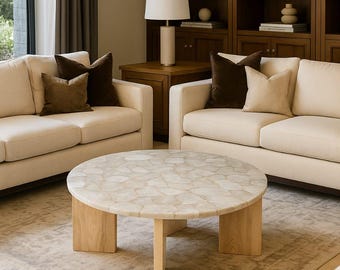 May include: A round coffee table featuring a mosaic-style top composed of irregular, light-coloured stone pieces. The table is supported by three thick, light-toned wooden legs arranged in a tripod configuration, providing a stable base.