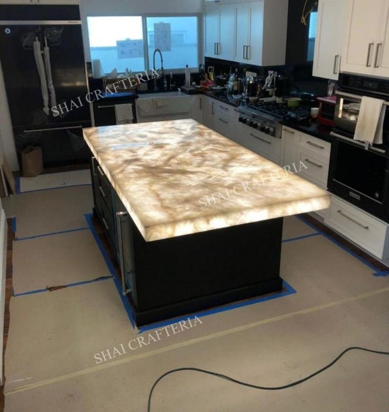 May include: A kitchen island with a glowing, light-colored countertop. The countertop has a marbled appearance with shades of beige and cream. The base of the island is black with drawers and cabinet doors. The kitchen has white cabinets and stainless steel appliances.