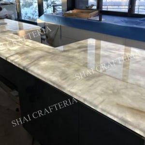 May include: An L-shaped countertop with a light-colored, veined surface. The countertop is supported by a dark base. The surface reflects light, and the words "SHAI CRAFTERIA" are visible on the side.