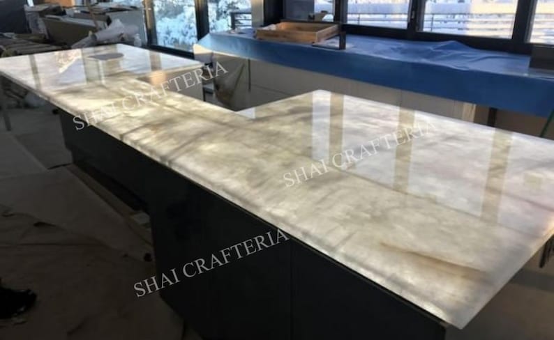 May include: An L-shaped countertop with a light-colored, veined surface. The countertop is supported by a dark base. The surface reflects light, and the words "SHAI CRAFTERIA" are visible on the side.