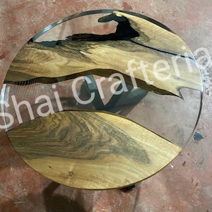 May include: A round table featuring a natural wood slab with prominent grain patterns embedded in clear resin. The wood has dark, irregular edges and light brown tones. The text Shai Crafteria is overlaid across the centre of the table surface.