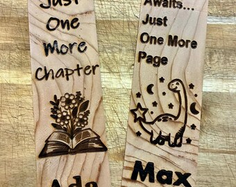 May include: Two wooden bookmarks with engraved text and designs. One reads "Just One More Chapter" with an open book and floral design, and the name "Ada". The other says "Adventure Awaits... Just One More Page" with a dinosaur and stars, and the name "Max".