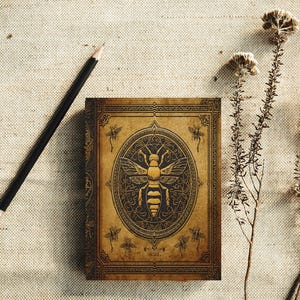 May include: A brown journal featuring a detailed bee design on the cover, with gold-coloured accents. A black pencil lies next to the journal, and dried plants are in the background. The journal has an antique aesthetic.