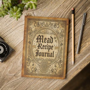 May include: A vintage-style "Mead Recipe Journal" with ornate border design and text. The journal is open on a wooden surface, next to a pencil and a pen. The cover is a tan colour with dark brown text and illustrations.